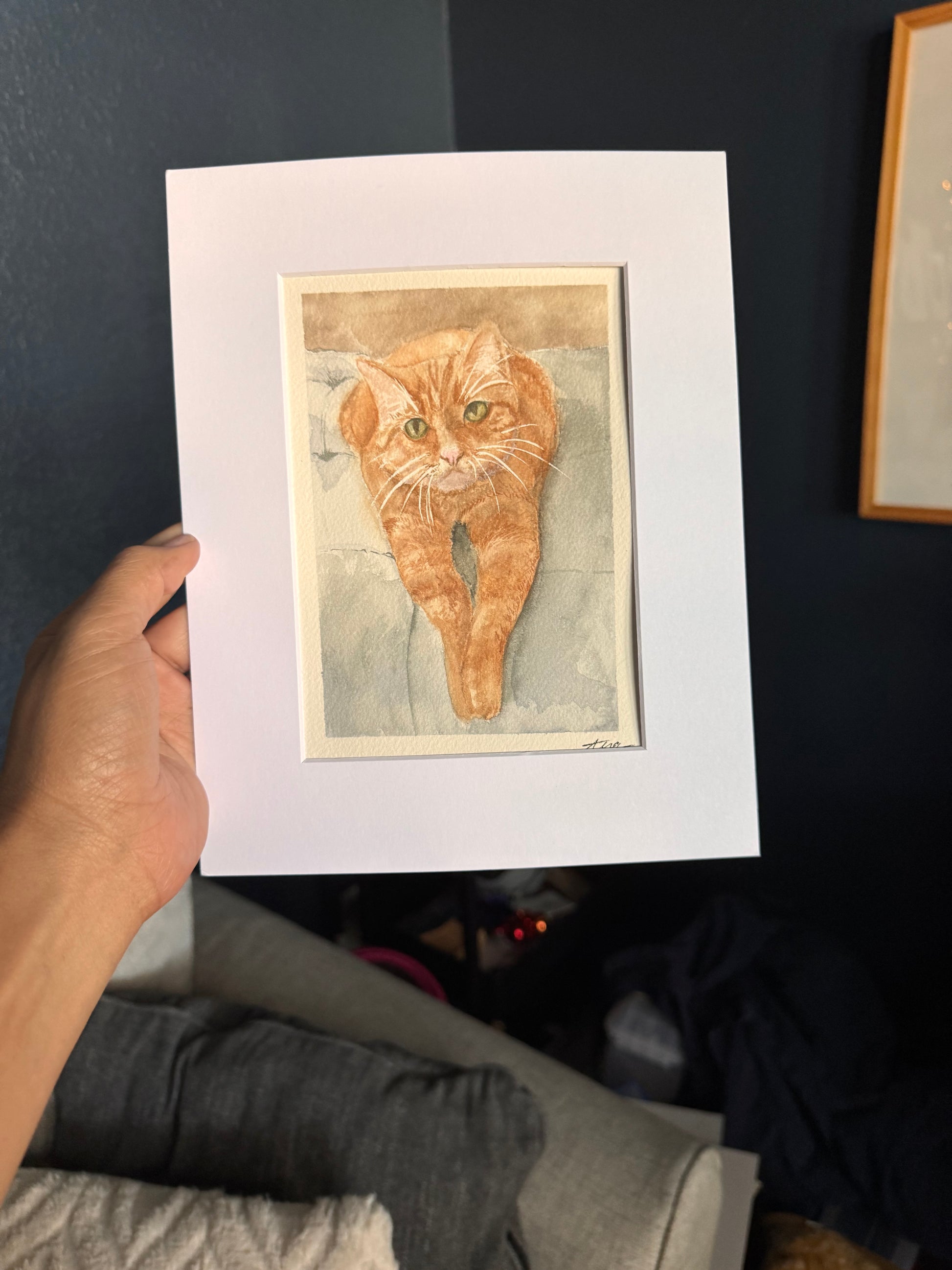Framed artwork of a cat held by a person on a dark surface.